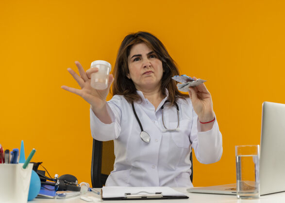 空着神经质的中年女医生 穿着医用长袍 带听诊器 坐在办公桌旁 用笔记本电脑和医疗工具 拿着空罐子和药片 背景是橙色 有复印空间笔记本电脑药片医疗