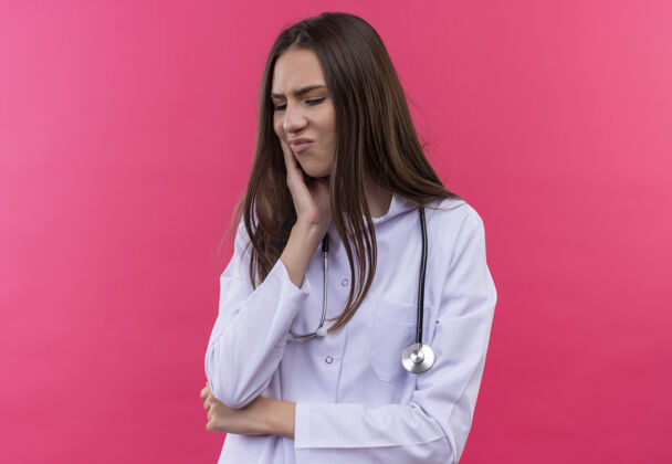 女孩生病的年轻女医生穿着听诊器医用长袍 把她的手放在孤立的粉红色背景上疼痛的牙齿上穿着疼痛手