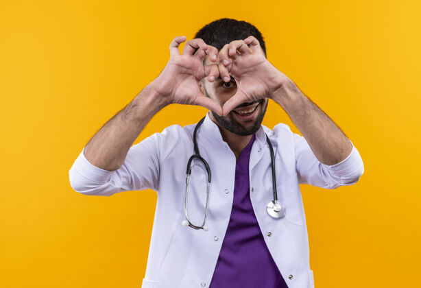 医生微笑着的年轻男医生穿着听诊器医用长袍 在孤立的黄色背景上显示心脏的姿势年轻手势礼服