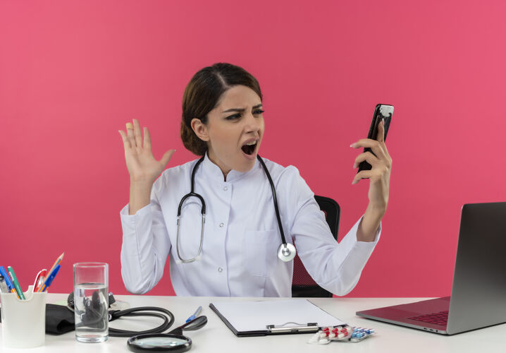 办公桌愤怒的年轻女医生穿着医用长袍 手持听诊器坐在办公桌旁 拿着医疗工具在电脑上工作 拿着手机 看着隔离的粉色墙壁上有复印空间愤怒年轻长袍