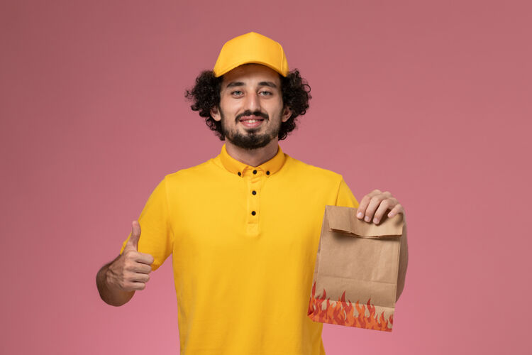 帽子正面图穿着黄色制服的男快递员拿着浅粉色墙上的纸质食品包制服手持快递员