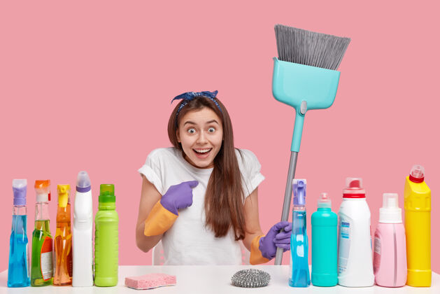 清洁惊奇快乐的年轻女子指着自己 在清洁服务工作 拿着扫帚 坐在桌子旁用洗涤剂和除臭剂扫帚清洁家务