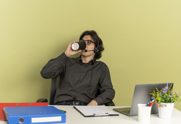 自信年轻自信的上班族戴着耳机戴着眼镜坐在办公桌旁 拿着办公工具用笔记本电脑喝着咖啡工具坐工作人员
