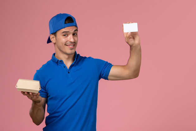 快递正面图：身穿蓝色制服披肩的年轻男性信使手拿着小快递食品包和浅粉色墙上的白色卡片工作人员包裹