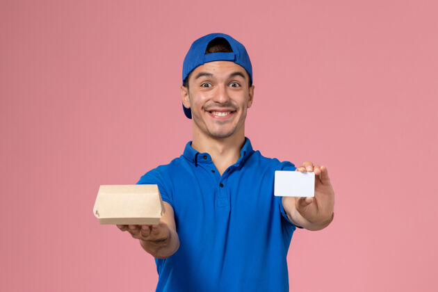 粉色正面图：身穿蓝色制服披肩的年轻男性信使手拿着小快递食品包和浅粉色墙上的白色卡片制服工作送货