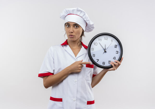 厨师惊讶的年轻女厨师穿着厨师制服拿着并指着墙上有抄写空间的钟惊讶穿着墙