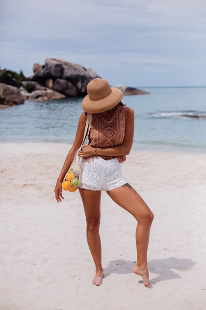 沙滩年轻漂亮的白种人 皮肤黝黑 穿着针织服装 戴着草帽 手拿一个装满水果 苹果 橘子 葡萄的线袋 在热带海滩上度假手提包女士