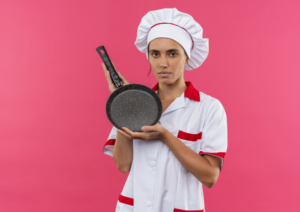 女年轻的女厨师穿着厨师制服 拿着煎锅盘站在隔离的粉色墙壁上 还有复印空间看着煎着年轻