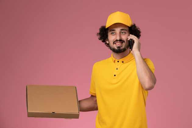 制服正面图身穿黄色制服的男快递员拿着食品快递箱 在浅粉色的墙上讲电话食物盒子电话