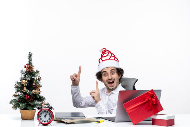 圣诞节新的一年的心情与惊讶的微笑兴奋的年轻商人坐在办公室里 并指着上面的白色背景成年人年轻商人