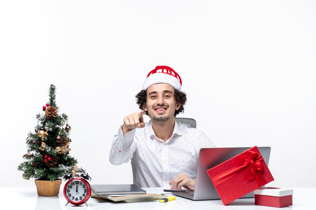 笔记本电脑满意的年轻商人与有趣的圣诞老人帽子指向某物 并在白色背景的办公室庆祝圣诞节满意微笑年轻商人