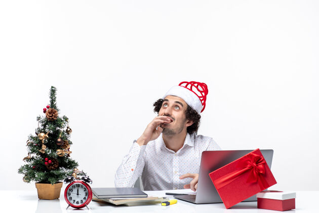满意满意积极的年轻商人 戴着有趣的圣诞老人帽 在白色背景的办公室里思考和庆祝圣诞节坐着人年轻的商人
