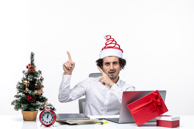 指着困惑的年轻商人 戴着滑稽的圣诞老人帽子 指着办公室左边白色背景上的东西成年人困惑笔记本电脑