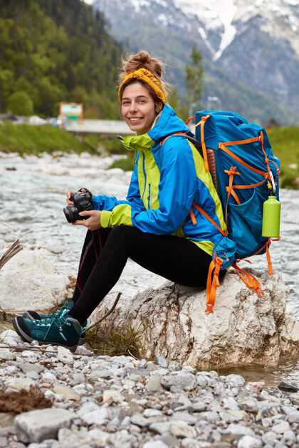 岩石疲惫的女游客坐在山上小溪边的石头上 手持专业相机 观看照片高兴冒险背包