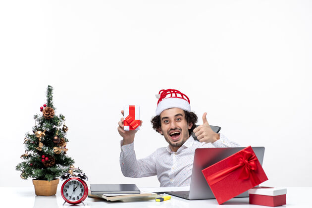 帽子带着微笑的新年心情满意的年轻商人戴着圣诞老人的帽子坐在办公室里 拿着白色背景的礼物年轻的商人坐着礼物