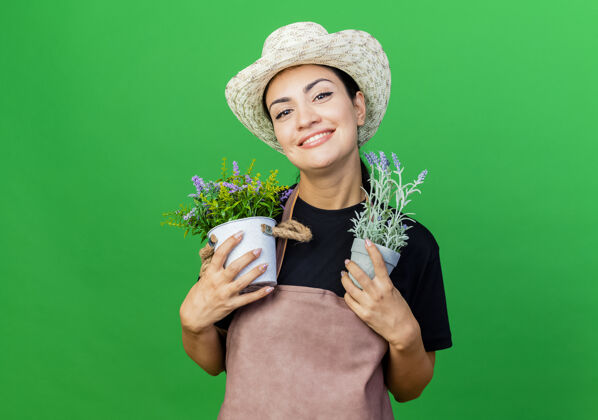 微笑年轻漂亮的女园丁围着围裙 戴着帽子 手里拿着盆栽植物 脸上洋溢着幸福的笑容植物年轻园丁