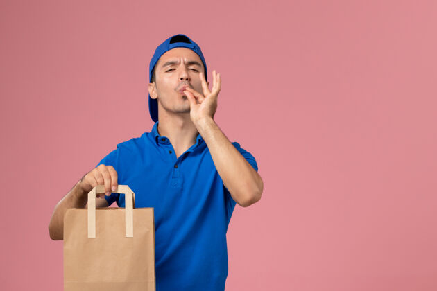 男性正面图：身穿蓝色制服和披风的年轻男性快递员 手上拿着一个纸包裹 放在粉红色的墙上年轻人成人工作