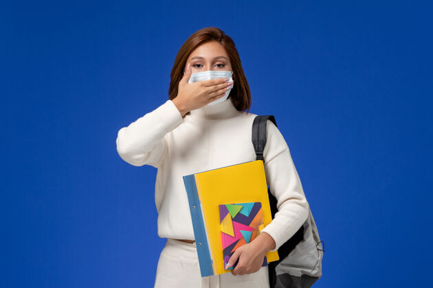 学生正面图身穿白色运动衫的年轻女学生戴着面具 手上拿着书包和文案 在蓝色的墙上闭着嘴穿戴风景文案