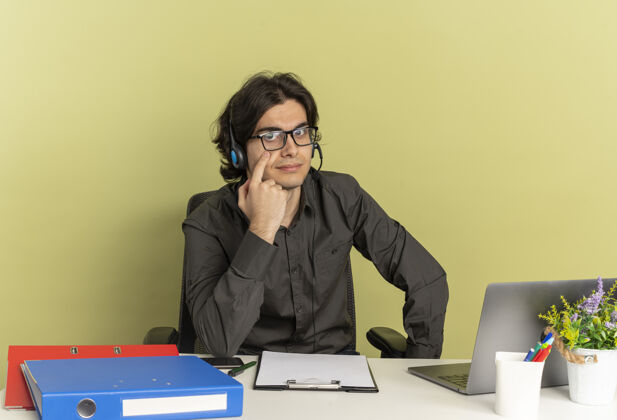 看年轻自信的上班族戴着耳机戴着眼镜坐在办公桌旁 拿着办公工具用笔记本电脑指着眼睛办公桌点笔记本电脑