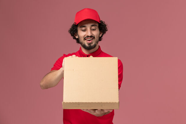 人正面图：身穿红色衬衫和披风的男性快递员手持浅粉色墙上的送货食品箱快递微笑视图