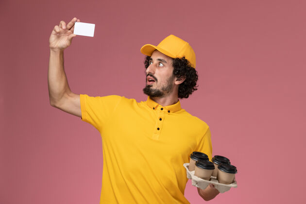 杯子正面图：身穿黄色制服的男信使手拿着棕色的咖啡杯和白色卡片放在粉红色的墙上帽子制服卡片