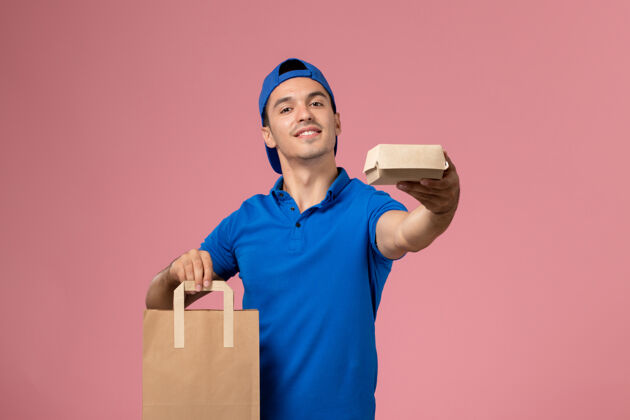快递正面图：身穿蓝色制服和披风的年轻男性快递员 手上拿着快递包裹 站在粉红色的墙上人制服视图