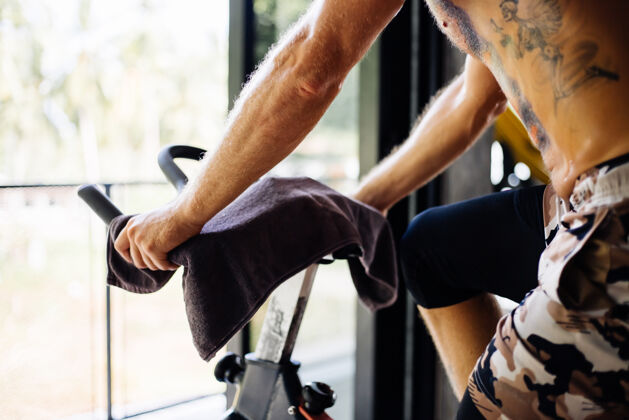 努力纹身 肌肉发达 胡须浓密的男人骑着自行车在体育馆里锻炼有氧运动 靠近大窗户 外面可以看到树骑自行车运动锻炼