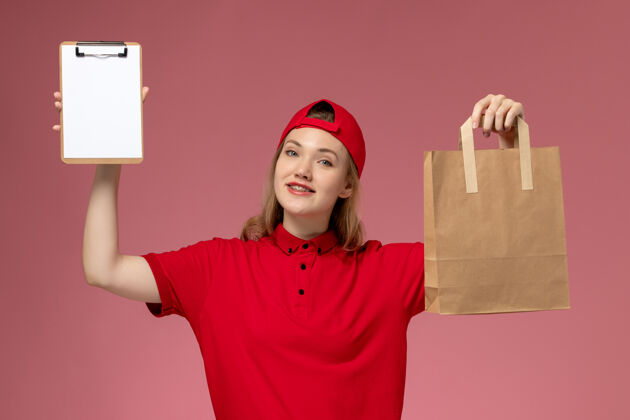 人正面图身穿红色制服的年轻女快递员拿着快递食品包和笔记本在浅粉色的墙上微笑快乐视图递送