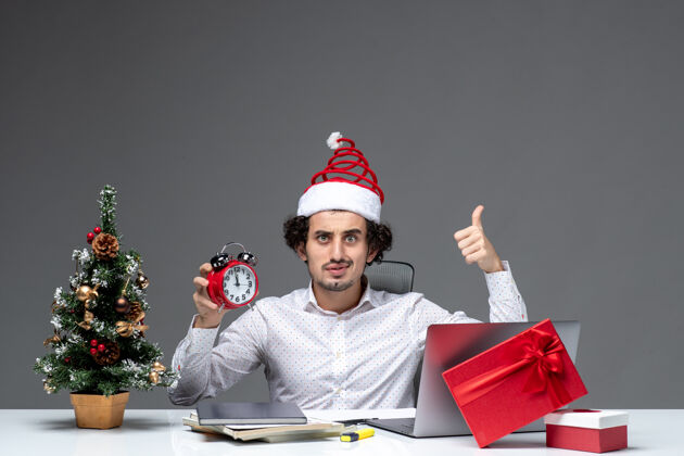 时钟年轻兴奋震惊的商人戴着圣诞老人的帽子 在黑暗的背景下独自一人坐在办公室里看时钟人成人圣诞老人