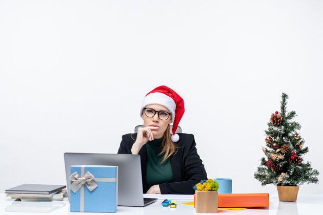 桌子一位严肃的商务女士 戴着圣诞老人帽 坐在一张桌子旁 桌子上放着圣诞树和礼物 她仔细地注视着白色背景上的东西礼物电脑笔记本电脑