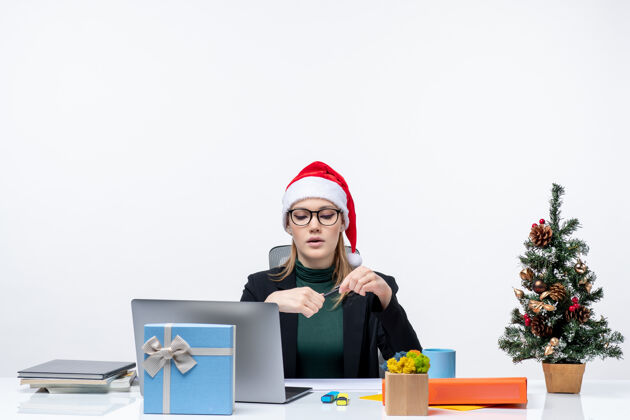 笔记本电脑新年心情严肃的金发女人戴着圣诞老人的帽子坐在一张桌子旁 桌子上放着一棵圣诞树和一份白色背景的礼物金发女人女人圣诞老人