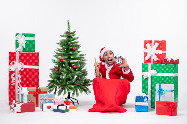 圣诞节好奇的年轻人坐在地上 拿着钟在礼物和圣诞树旁庆祝新年或圣诞节地男人礼物