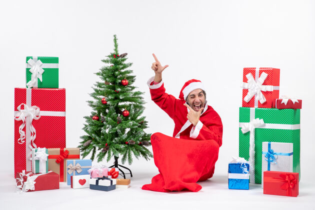 年轻人有趣的情感兴奋的年轻人打扮成圣诞老人与礼物和装饰圣诞树坐在地上指着上面的白色背景礼物成人年轻人
