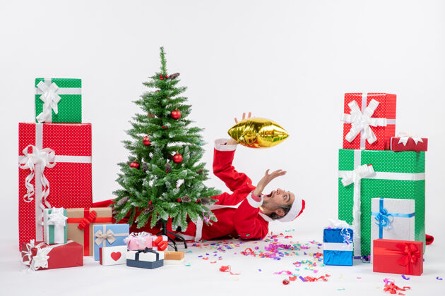 圣诞老人圣诞气氛与年轻的圣诞老人躺在圣诞树后 在白色背景上不同颜色的礼物附近季节说谎礼物