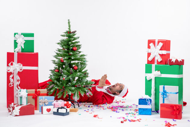 冬青节日气氛与年轻震惊圣诞老人躺在圣诞树后靠近礼物白色背景季节年轻圣诞老人