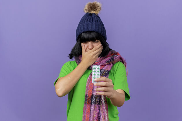 疼痛疼痛的年轻白种人 戴着冬天的帽子 戴着围巾 手里拿着一包医药胶囊 手放在嘴上 闭着眼睛 忍受着牙痛围巾胶囊穿着