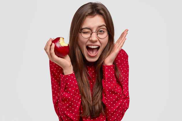 维生素高兴的年轻女子头像眨着眼睛 举手靠近头部 咬着新鲜的苹果 有喜悦的表情 穿着红色圆点衬衫眼镜高兴女性