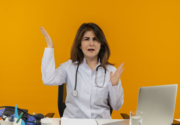 坐着困惑的中年女医生穿着医用长袍 手持听诊器 坐在办公桌旁 手持医疗工具 在隔离的橙色墙壁上举手 还有复印空间工具工作困惑