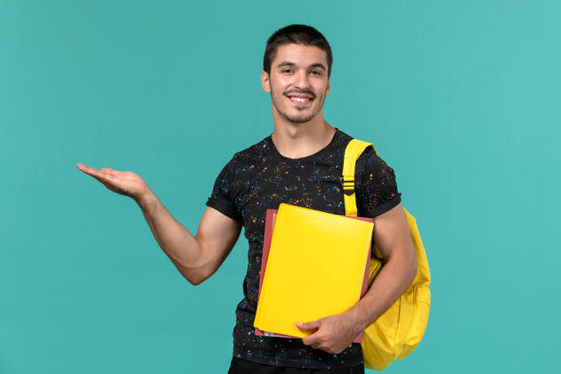 学生身穿深色t恤黄色背包的男学生正面图 浅蓝色墙上放着不同的文件便携式电脑个人男学生