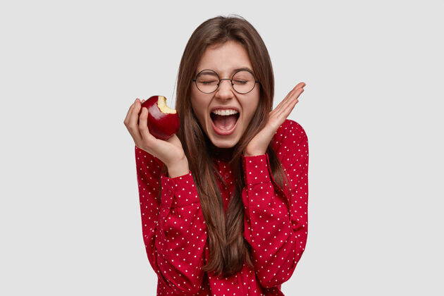 饮食快乐的黑发女士积极地喊叫和手势 咬新鲜的苹果 闭上眼睛 享受健康的饮食眼镜服装凝视
