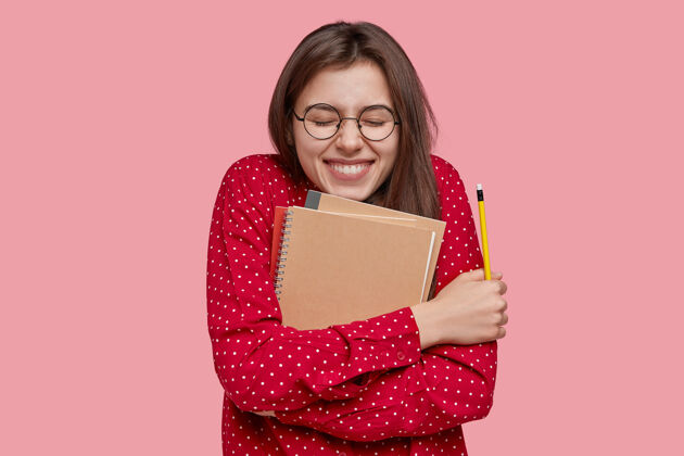 高兴表情积极的女生随身携带笔记本 笑容灿烂 手持铅笔 穿着时尚的红衬衫 隔离在粉色背景上教育满意学生