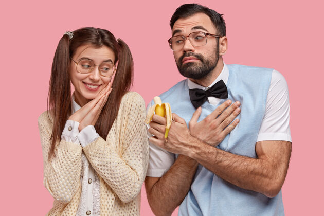 惊讶体贴的男友留着厚厚的胡茬 穿着优雅 看起来很尴尬 手放在胸前 建议女友咬香蕉美味水果表达