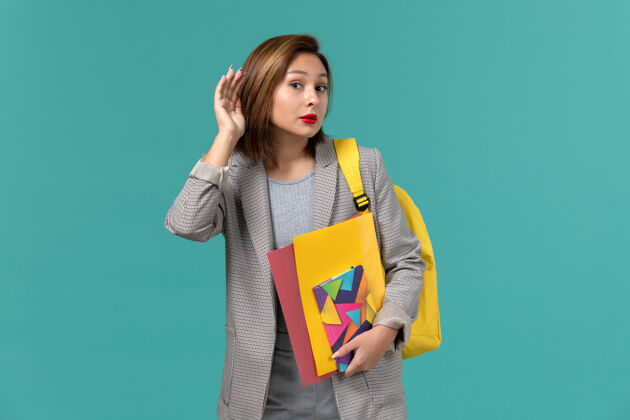 大学身穿灰色夹克 背着黄色背包 拿着文件和文案的女学生正试图在蓝色的墙上听到声音女学生快乐听到