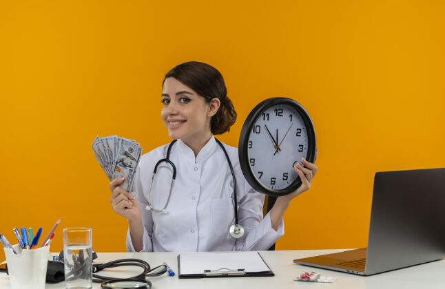 听诊器面带微笑的年轻女医生穿着医用长袍 手持听诊器坐在办公桌旁 拿着医疗工具 拿着挂钟和隔离黄色背景上的现金坐墙壁医生