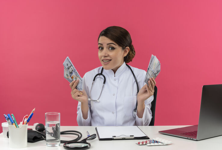 年轻人年轻的女医生穿着医用长袍 手持听诊器 坐在办公桌旁 拿着医疗工具 拿着现金 站在隔离的粉色墙上 还有复印空间坐着听诊器女