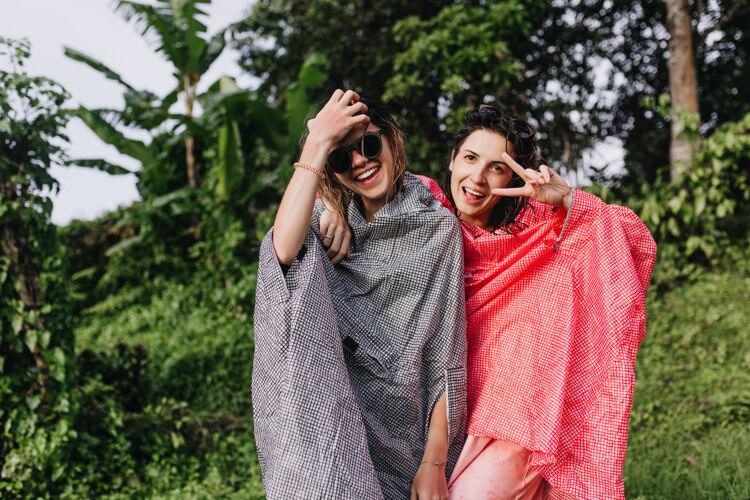 女人穿着粉红色雨衣的兴奋的黑发女人和最好的朋友玩得很开心可爱的姐妹们在异国森林里度过时光的户外照片天气巴厘岛探险