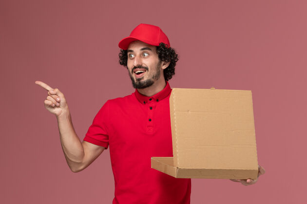 送货正面图：身穿红色衬衫和披风的男性快递员手持浅粉色墙上的送货食品箱男性快递披萨员工