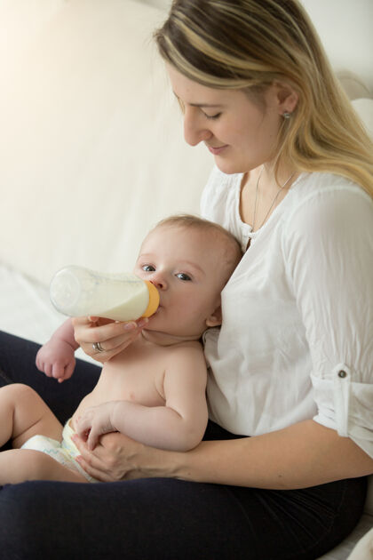 母性年轻母亲坐在床上给婴儿喂奶瓶的色调肖像渴可爱妈妈
