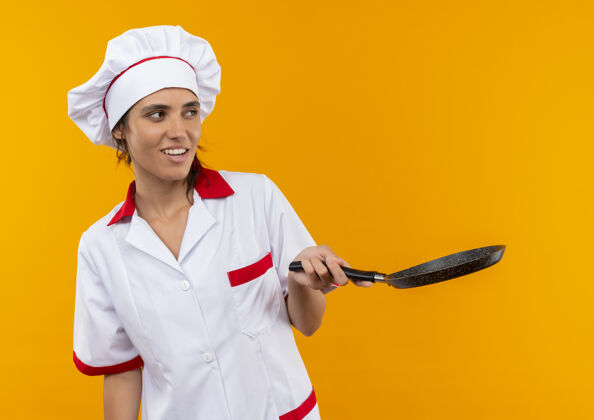拿着看着旁边穿着厨师制服的年轻女厨师拿着煎锅站在隔离的黄色墙壁上复制空间边平底锅制服
