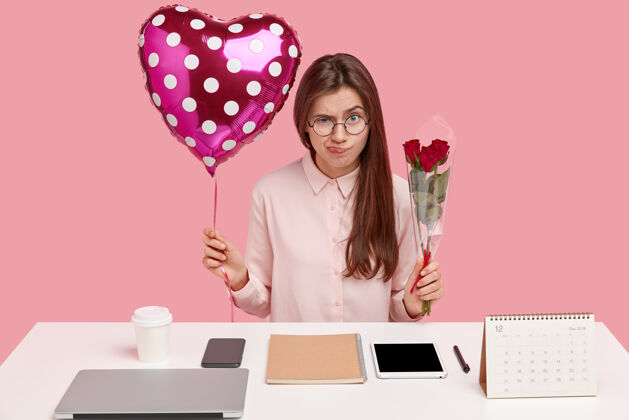 气球照片中的黑发女士不满地皱起眉头 拿着情人节礼物和玫瑰花束 心烦意乱地收到陌生人的礼物 拿着笔记本摆在桌面上电脑礼物女性